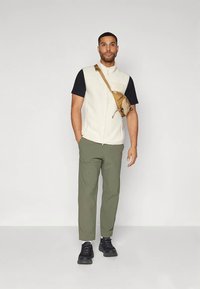 Άνδρας στέκεται φορώντας ένα λευκό αμάνικο μπλουζάκι πάνω από ένα μαύρο πουκάμισο, πράσινα παντελόνια, μαύρα αθλητικά παπούτσια και μια καφέ τσάντα χιαστί.