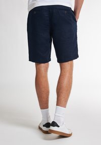 Pantaloni corti blu navy con vestibilità rilassata, realizzati in tessuto testurizzato. Presentano due tasche posteriori con chiusura a bottone. Indossati con calzini bianchi e scarpe.