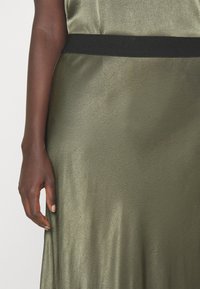 Abito in satin verde oliva con una texture liscia, dotato di una vita nera. L'immagine mostra una mano che tiene il tessuto di lato.