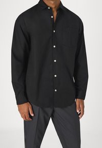 Man i en svart långärmad skjorta med knappar och ficka på bröstet, som är ihopkopplad med mörkgrå mönstrade byxor.