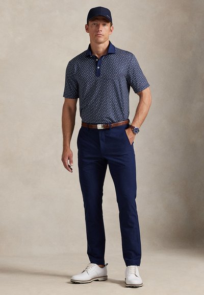 Μπλε σκούρο πόλο με σχέδιο και σκούρο μπλε γιακά, συνδυασμένο με μπλε σκούρο chino παντελόνια και λευκά παπούτσια. Το μοντέλο φορά μπλε σκούρο καπέλο και καφέ ζώνη.