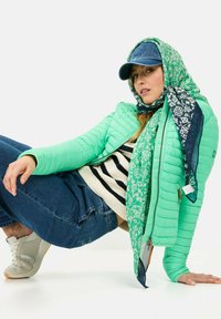 Grøn polstret jakke med quiltet tekstur, parret med en stribet sweater og blå denimjeans. Tilbehøret inkluderer et mønstrede tørklæde og en kasket.