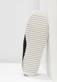 Chaussure en toile noir avec une semelle en caoutchouc blanc texturé dotée de rainures horizontales et d'une finition en fibres naturelles sur le bord.