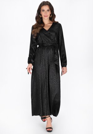 Hosszú hullámos hajú nő fekete, leopárdmintás, hosszú ujjú átöltős ruhában és fekete magassarkú szandálban, előre sétálva.