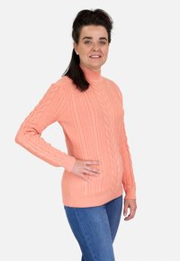 Suéter de punto trenzado de color durazno con un cuello alto, puños y dobladillo acanalados. Lleva puesto sobre jeans azules. Textura suave, mangas largas.