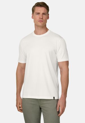 Homme portant un t-shirt blanc uni à manches courtes et un pantalon gris, debout devant un fond blanc.