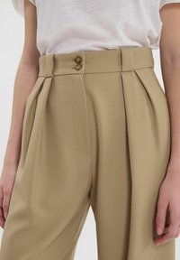 Pantalon beige taille haute avec fermeture à double bouton, plis et poches latérales. Le tissu présente une texture subtile.