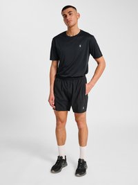 Młody mężczyzna stojący, ubrany w czarne sportowe szorty i koszulkę, białe skarpety i czarne sneakersy, ręka w kieszeni, neutralne tło.