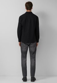 Schwarzes langärmliges Hemd mit geradem Schnitt, geknöpften Manschetten und einem Rückeneinsatz, kombiniert mit dunkelgrauen Jeans und schwarzen Schuhen.