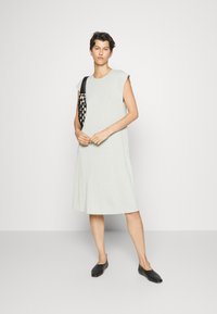 Vestido verde claro, de mangas curtas, feito de um tecido suave, apresentando um corte solto e bolsos laterais. Combinado com sapatos pretos tipo slip-on e uma mala estampada.