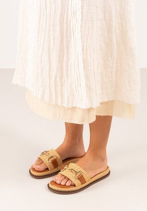 Sandalias tejidas marrones con dos correas, con acentos de hardware dorado y una suela texturizada, combinadas con un vestido blanco en capas.
