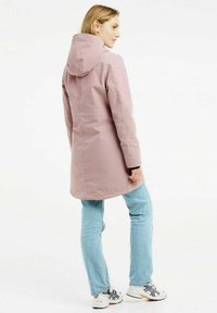Manteau imperméable rose clair avec capuche, présentant une coupe droite, des manches longues et des finitions noires. Porté avec un jean bleu et des chaussures de sport.
