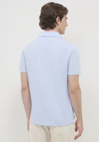 Un jeune homme avec des cheveux bruns bouclés portant un polo à manches courtes bleu clair et un pantalon beige, tourné le dos contre un fond uni.