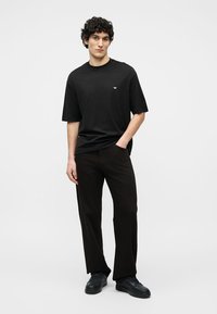 Νεαρός άνδρας με σγουρά μαλλιά που φοράει μια μαύρη φαρδιά μπλούζα, μαύρο παντελόνι φαρδύ στο κάτω μέρος και μαύρα αθλητικά παπούτσια, στέκεται με το ένα χέρι στην τσέπη.