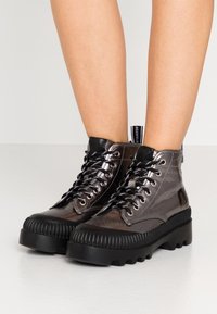Botas altas de color gris metálico con suelas de goma negras gruesas y cordones en blanco y negro, llevadas sobre piernas desnudas.