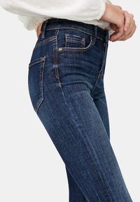 Jeans ajustados en denim azul oscuro con desvanecidos sutiles, que cuentan con un diseño clásico de cinco bolsillos y detalles de costura en dorado contrastante.
