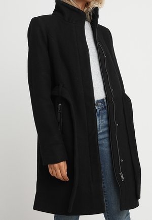 Frau trägt einen langen schwarzen Mantel mit hohem Kragen, offen, über einem hellgrauen Oberteil und blauen Jeans, stehend vor einem einfarbigen Hintergrund.