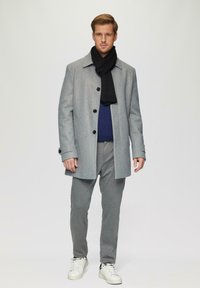 Grijze wollen jas met zwarte knopen, gecombineerd met een zwarte sjaal, blauwe trui, grijze broek en witte sneakers. Eenvoudig, modern ontwerp.