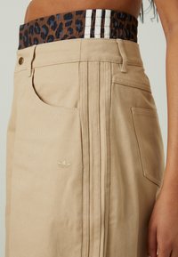 Beige Hose mit strukturierter Oberfläche, mit einem elastischen Bund im Leopardenmuster und drei weißen Streifen an der Seite. Besticktes Logo-Detail.