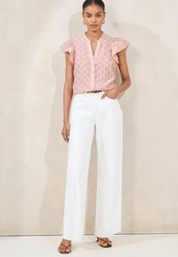 Blusa rosa a maniche corte con motivo testurizzato e bottoni, abbinata a pantaloni larghi bianchi. Il modello si trova in piedi con una mano nella tasca.