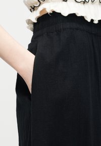 Χέρι στην τσέπη μαύρων παντελονιών με ελαστική μέση, φορεμένα με λευκό τοπ με βολάν και μαύρη επένδυση.