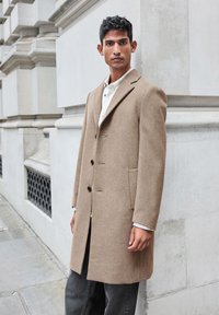 Cappotto in lana marrone chiaro con una silhouette sartoriale, design a doppio petto, rever a punta e tasche laterali. Chiusure con bottoni e dettagli di cucitura sottili.