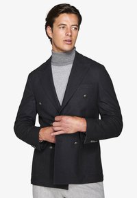 Sort dobbeltradet blazer med et struktureret stof, med spidse klapper og sølvfarvede knapper, lagd over en grå turtleneck top.