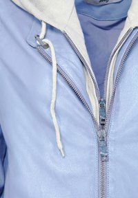 Vaaleansininen nahkatakki, jossa on kermahuppari, etuvetoketju ja nyörit. Sileä pinta, kaksivärinen muotoilu ja hillitty kiilto.