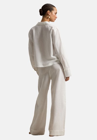 Vrouw met opgestoken haar, gekleed in een wit shirt met lange mouwen en broek met wijde pijpen, staand en zijwaarts kijkend tegen een effen achtergrond.