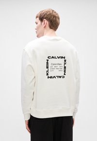 Λευκή μπλούζα με κουκούλα με εκτύπωση γραφικών στο πίσω μέρος που αναγράφει "Calvin Klein" και μια διεύθυνση με παχιά μαύρα γράμματα. Απλός, χαλαρός σχεδιασμός.