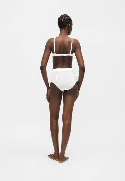Γυναίκα με πλεξούδες στα μαλλιά στέκεται ξυπόλητη με την πλάτη γυρισμένη, φοράει λευκά εσώρουχα με ψηλή μέση και ασορτί σουτιέν σε απλό γκρι φόντο.