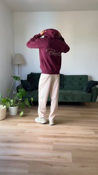 Moleton bordo com logo branco nas costas, combinado com calças de fato bege claro e sapatilhas brancas. Ambiente interior com sofá verde e planta.