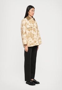 Bēšā ziedu žakete ar lielu apkakli, divām priekšējām kabatām un platām piedurknēm, saskaņota ar melnām taisnēm biksēm un melnām kurpēm.