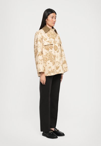 Beige kukka-ainetta oleva takki, jossa on suuri kaulus, kaksi etutaskua ja leveät hihat, yhdistetty mustiin suoriin housuihin ja mustiin kenkiin.