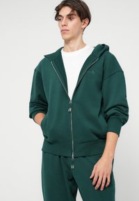 Zelená mikina na zip vyrobená z jemného materiálu, s kapucí, klokankovým kapsou a žebrovanými manžetami. Nositá přes bílý tričko.