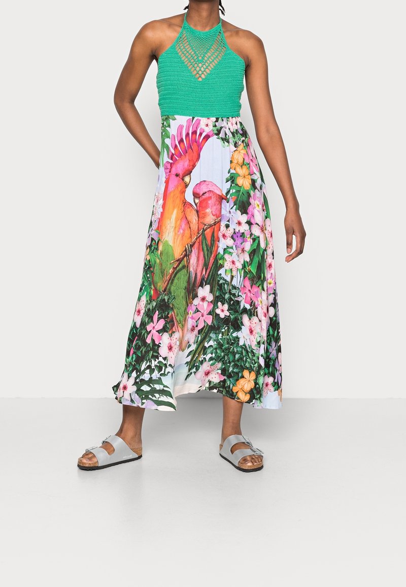 Femme portant un top dos nu en crochet vert et une jupe longue fleurie avec un grand imprimé perroquet coloré, associée à des sandales blanches.