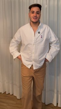 Giovane uomo con capelli corti e scuri in piedi con le mani sui fianchi, indossa una camicia bianca abbottonata e pantaloni beige davanti a tende bianche.