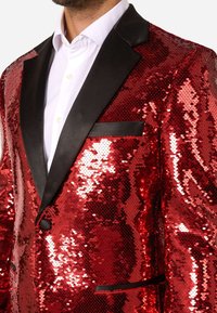 Sarkans spīdošs žakets ar melniem satīna ārmalēm un kabatu. Ar spīdīgām, pārklājošām spuldzītēm, kas rada teksturētu virsmu.