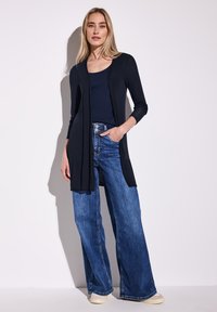 Largo cárdigan negro sobre una blusa azul marino combinado con jeans de pierna ancha de cintura alta en azul y zapatillas color crema. Tela suave con un diseño fluido.