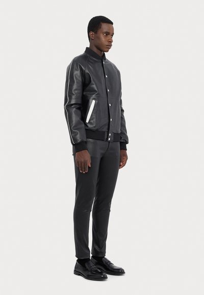 Black leather bomber jacket with ribbed cuffs, featuring a white zippered pocket and sleek, fitted black pants and shoes.