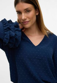 Top blu navy con scollo a V e pattern a pois testurizzati. Presenta maniche voluminose a balze con dettagli sottili. Texture del tessuto morbida.