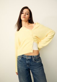 Cardigan jaune en maille avec ourlet côtelé, détails à boutons, porté sur un haut blanc et associé à un jean bleu taille haute avec des accents métalliques.