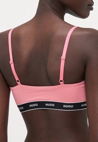 Soutien-gorge en tissu extensible rose avec bretelles ajustables. Bande noire avec le logo "HUGO" à la base. Design simple, texture lisse.