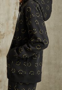 Black hoodie made of soft fabric, featuring an all-over pattern of yellow smiley faces. Includes a front pocket and adjustable hood.