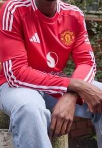 Červený dres Manchester United s bílými pruhy na rukávech a logem. Stylizováno se světle modrými džínami, sedící na strukturovaném povrchu.