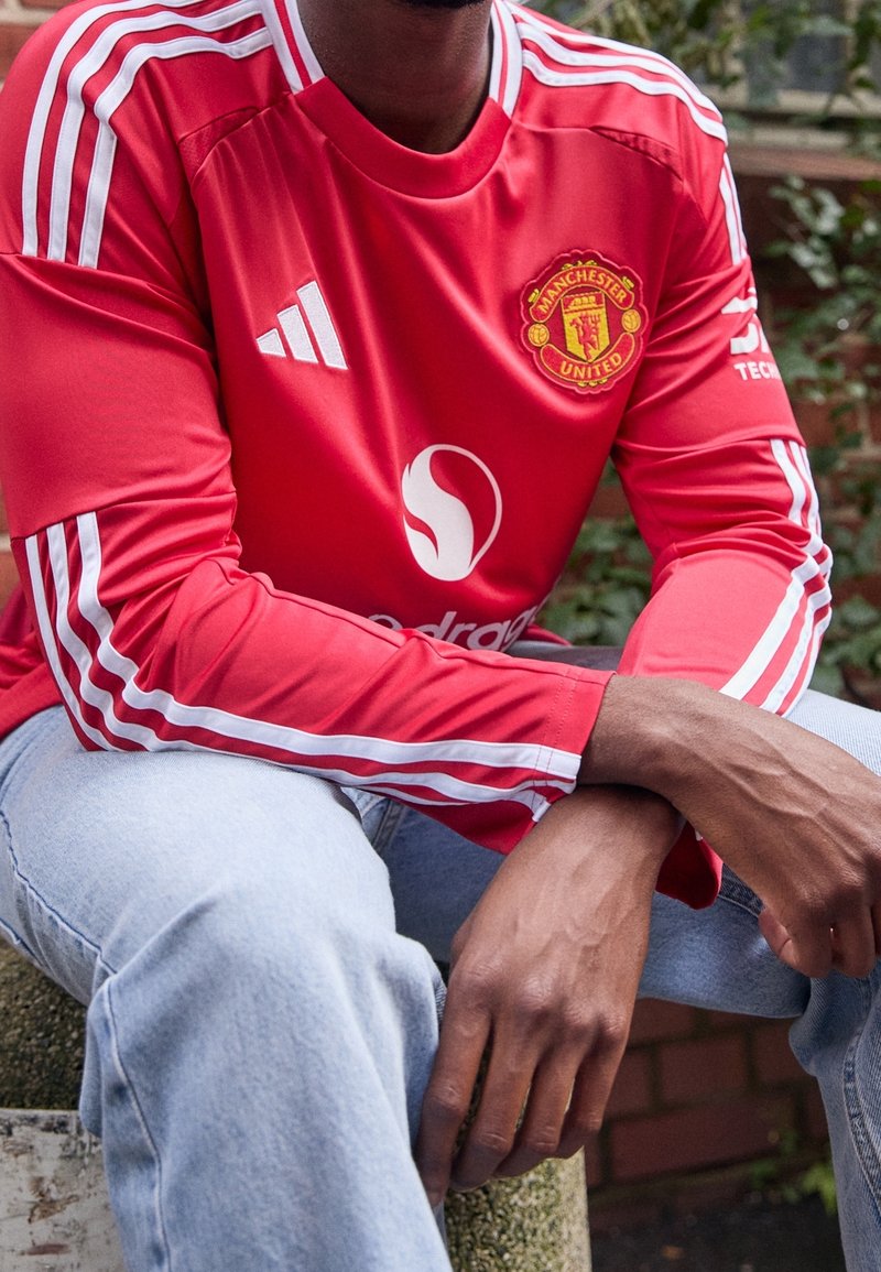 Červený dres Manchester United s bílými pruhy na rukávech a logem. Stylizováno se světle modrými džínami, sedící na strukturovaném povrchu.