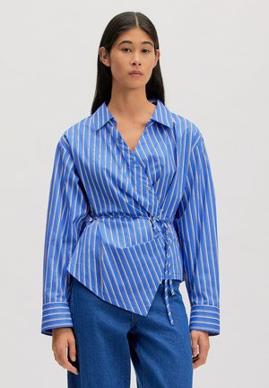 Frau mit langen dunklen Haaren, die ein blau-weiß vertikal gestreiftes Wickelhemd mit hochgekrempelten Ärmeln und blaue Jeans trägt.