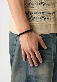 Juodas pintas odinis apyrankė, uždėta ant rankos, remiasi ant džinsų, su tekstūruotu raštu. Fonas neutralus.