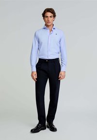 Camicia azzurro chiaro a maniche lunghe con colletto a camicia, emblema sul petto, abbinata a pantaloni scuri su misura e scarpe eleganti nere.