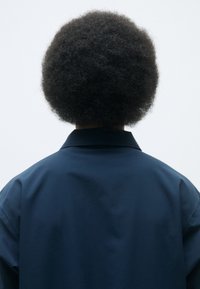 Marineblå, oversized jakke med en glatt tekstur og flat krage, som har en avslappet passform. Håret er stylet i en rund afroform.